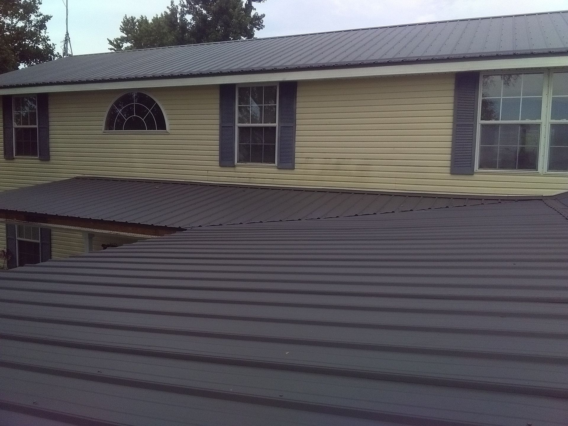 Residential building with beige siding and dark metal roof, seen from a lower rooftop level