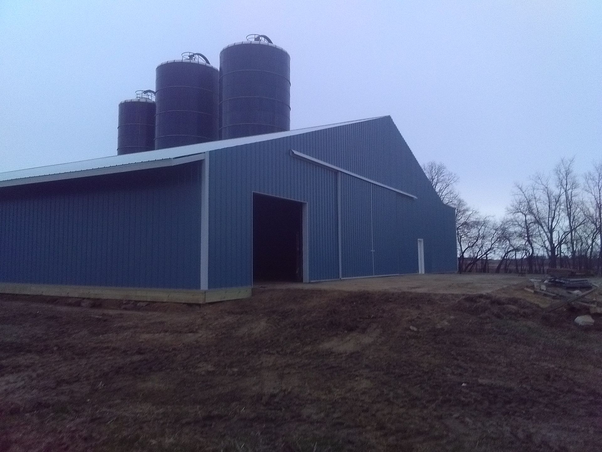 Blue barn with three silos on a dirt lot under an overcast sky