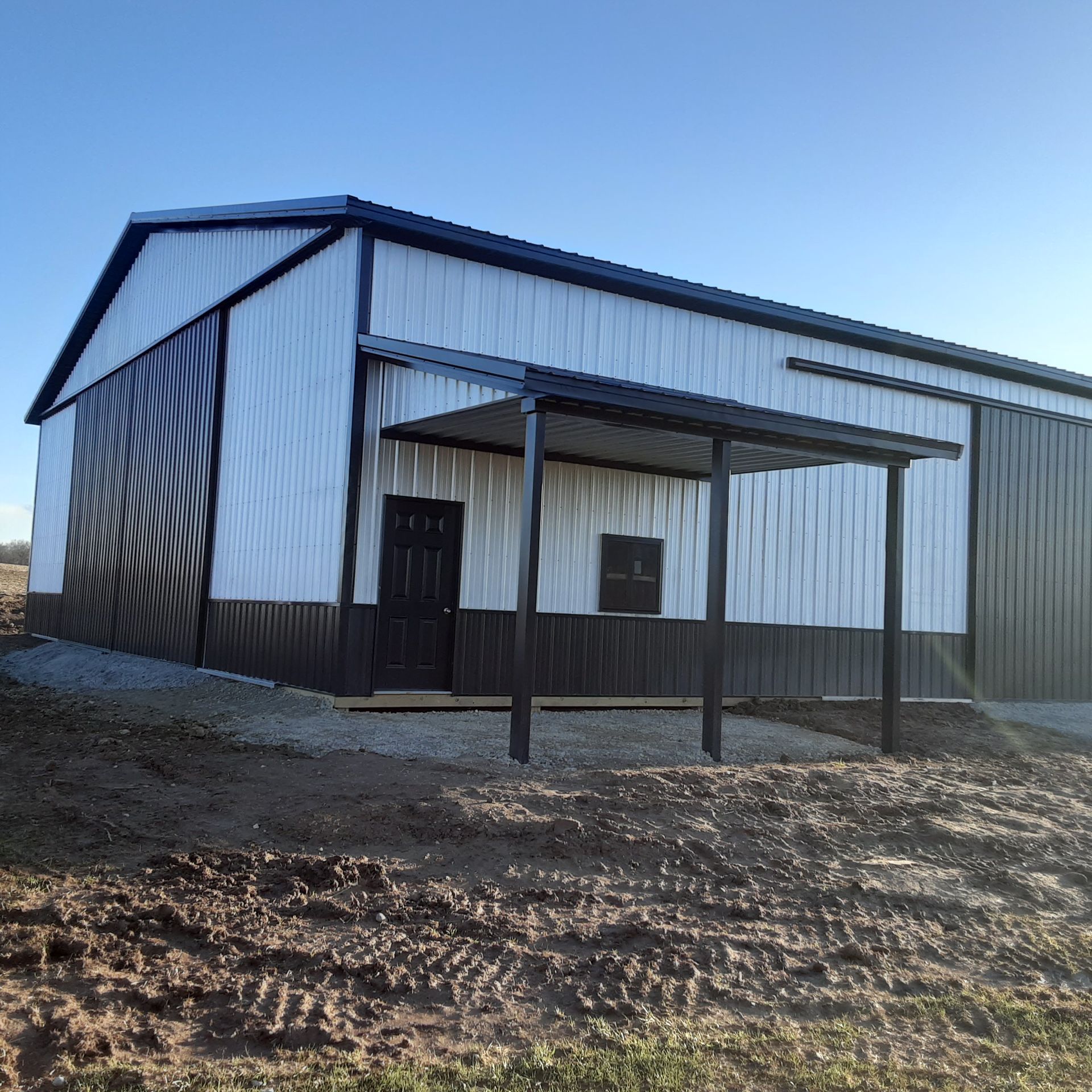 White metal barn with a small covered porch on a dirt lot under a clear blue sky.