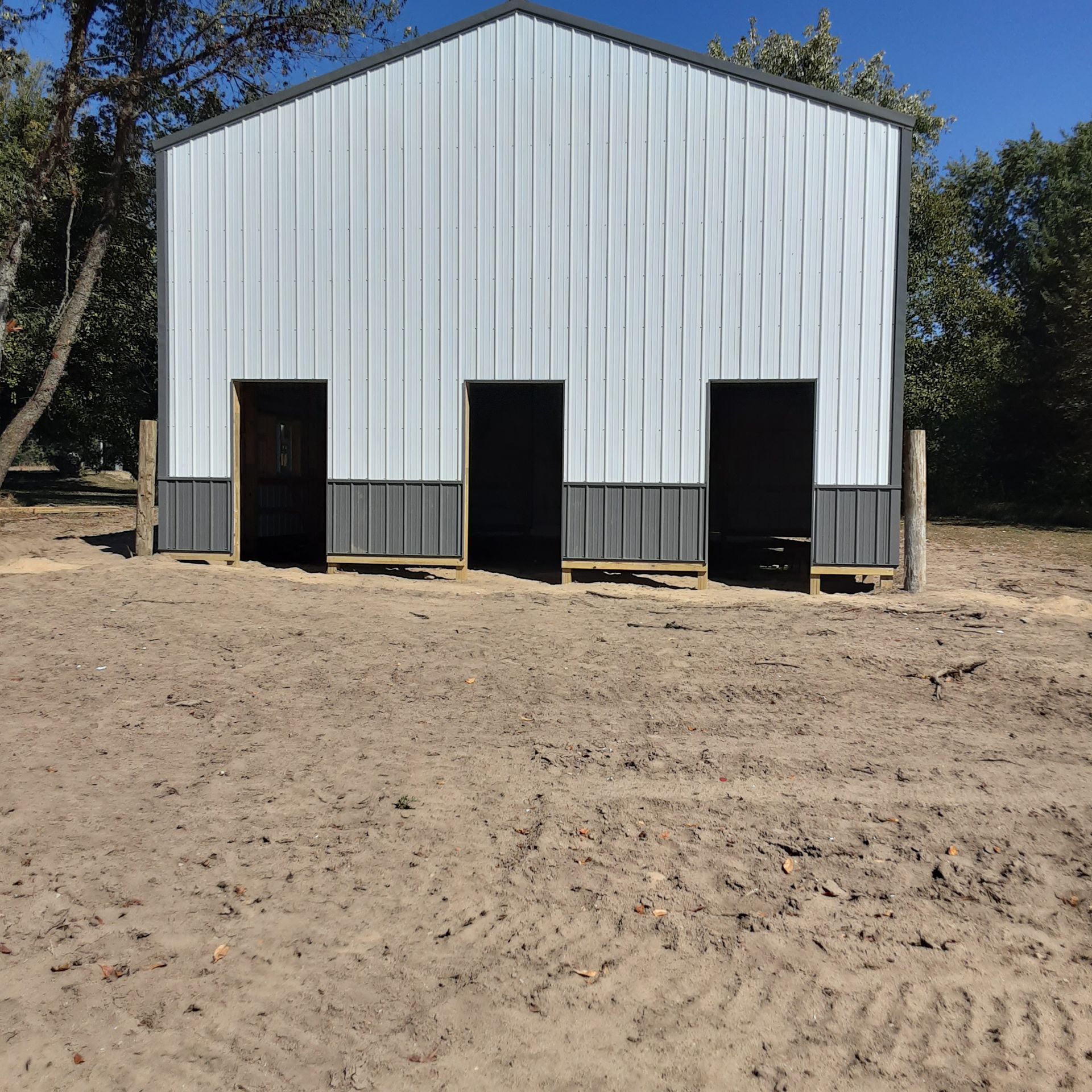 Small white metal building with three open doorways on a bare dirt lot, surrounded by trees.