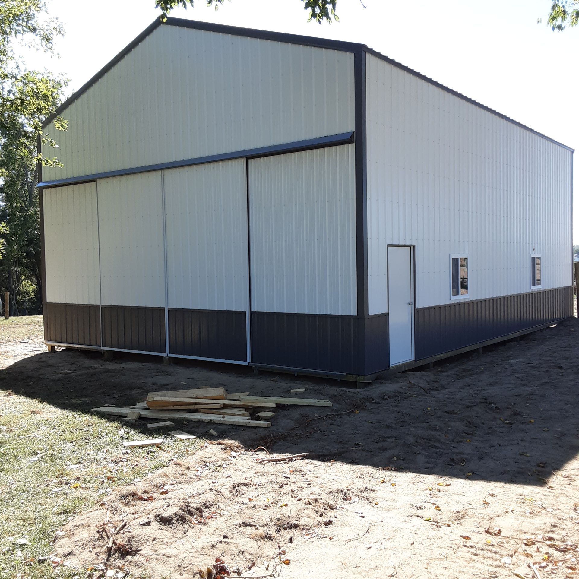 White metal shed with a dark base and a small side door, sitting on bare ground outdoors.