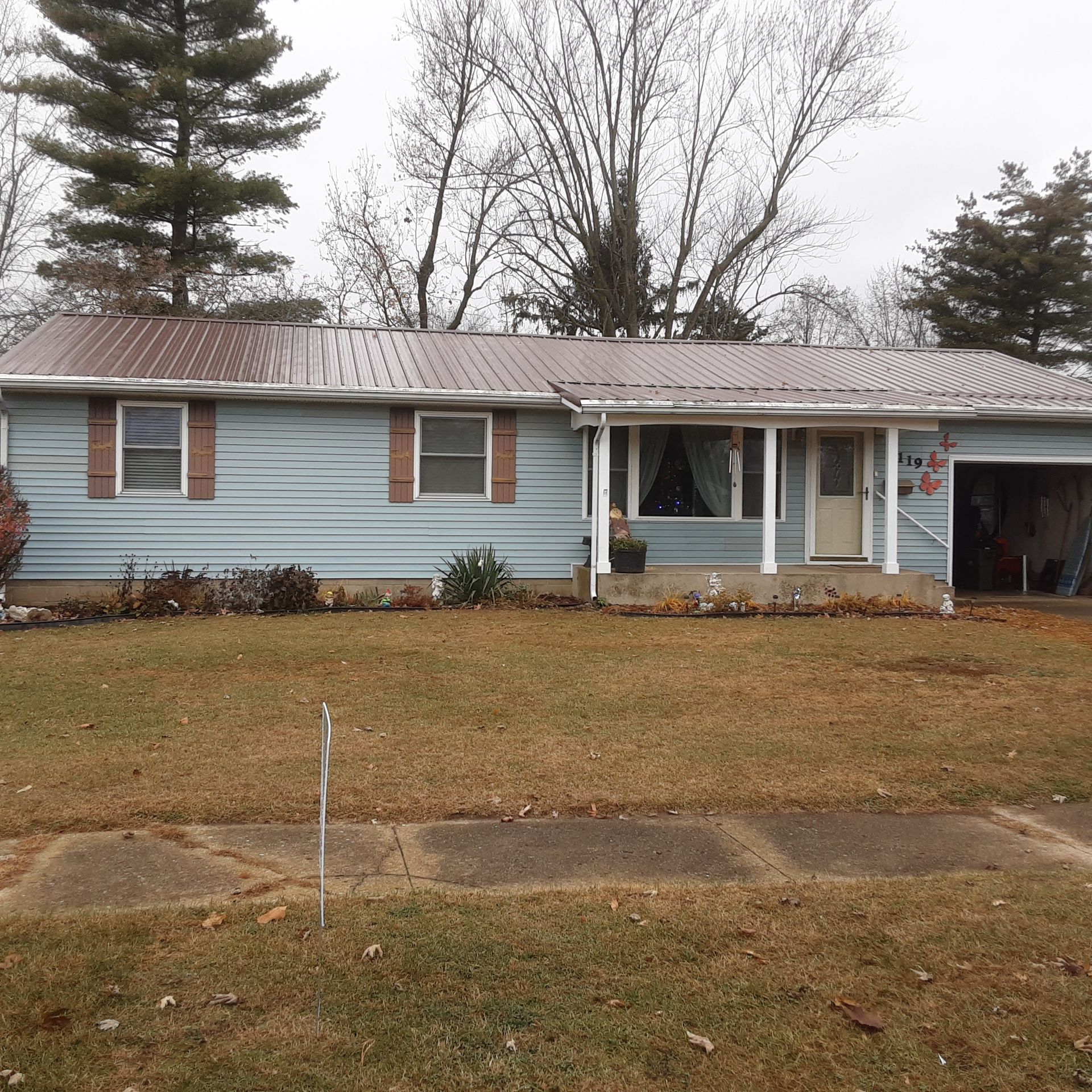 Single-story light blue house with a front porch, bare trees, and a brown lawn on an overcast day