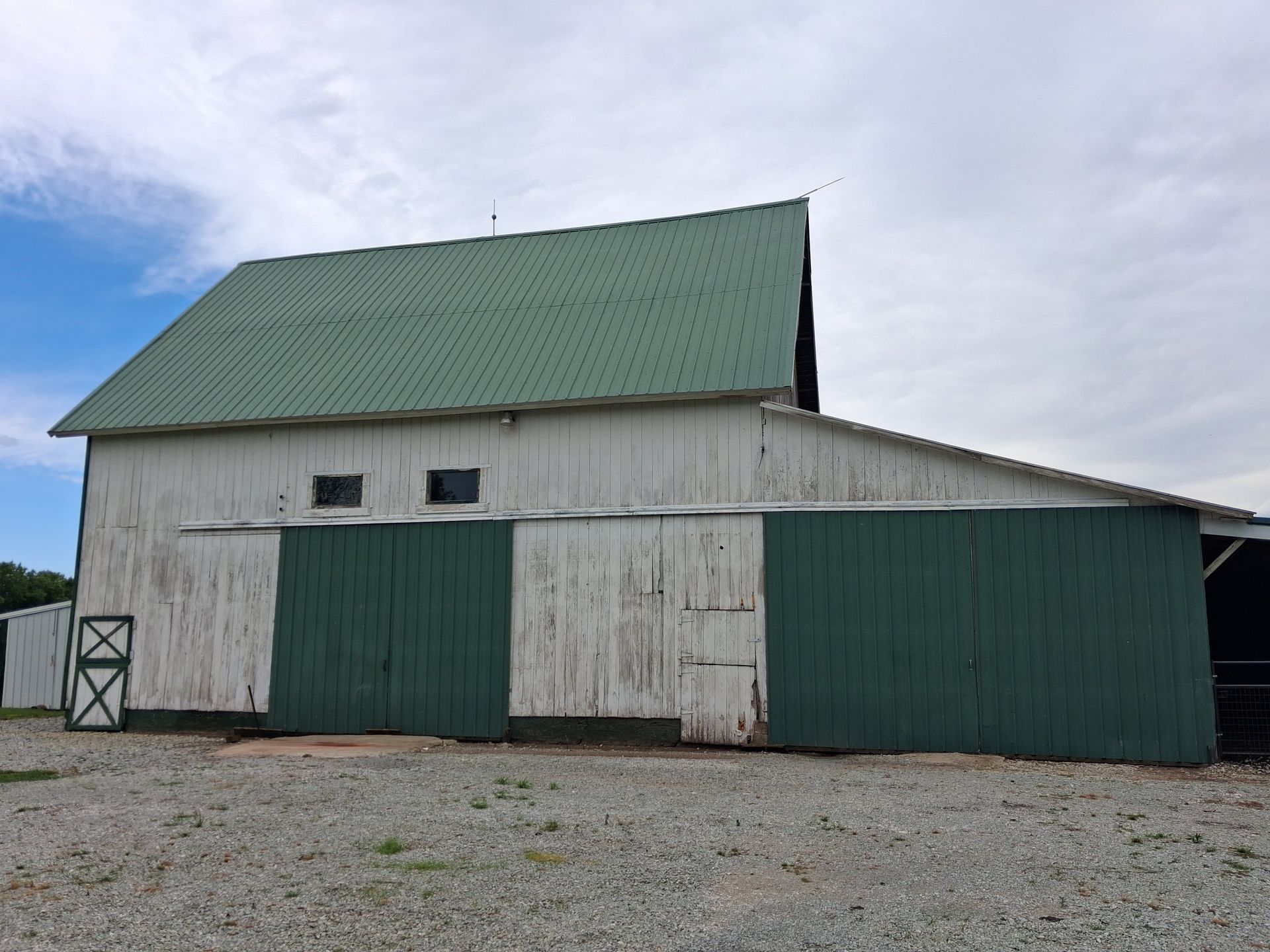 Weathered white barn with green roof and doors on gravel under a cloudy sky
