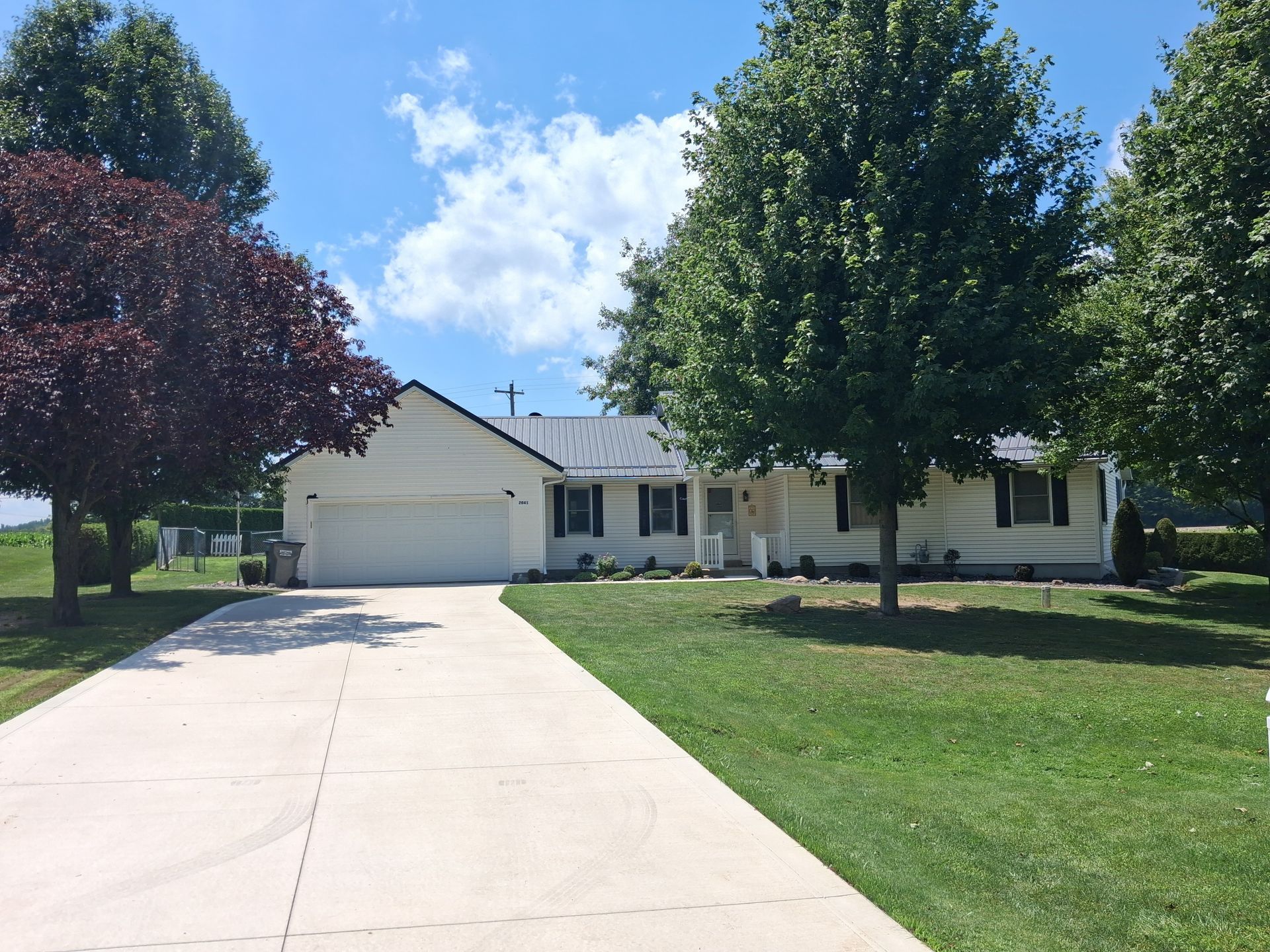 Suburban house with white siding, long driveway, and two trees on a sunny day