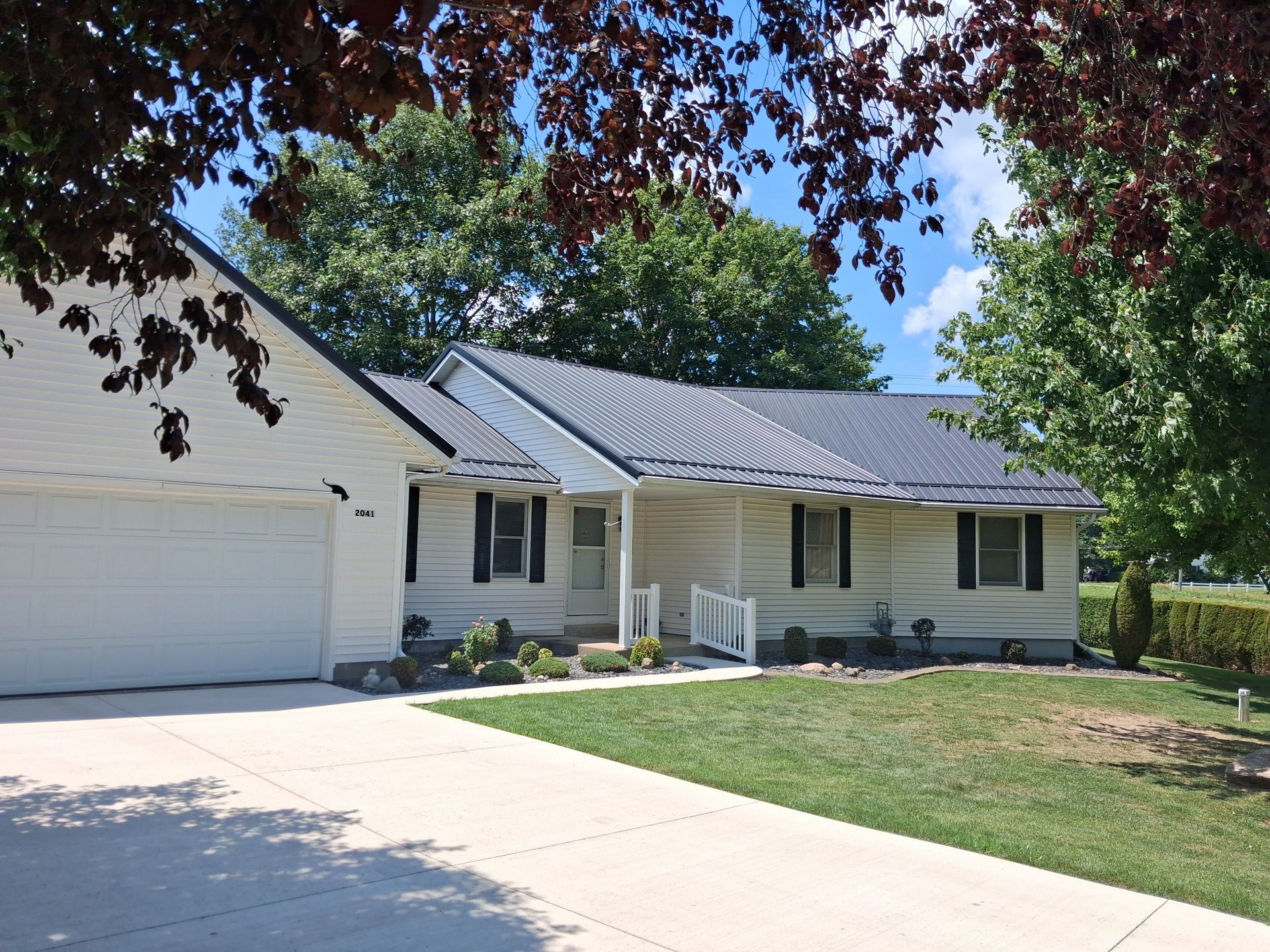 Single-story white house with gray roof, front porch, driveway, and green lawn under tree branches