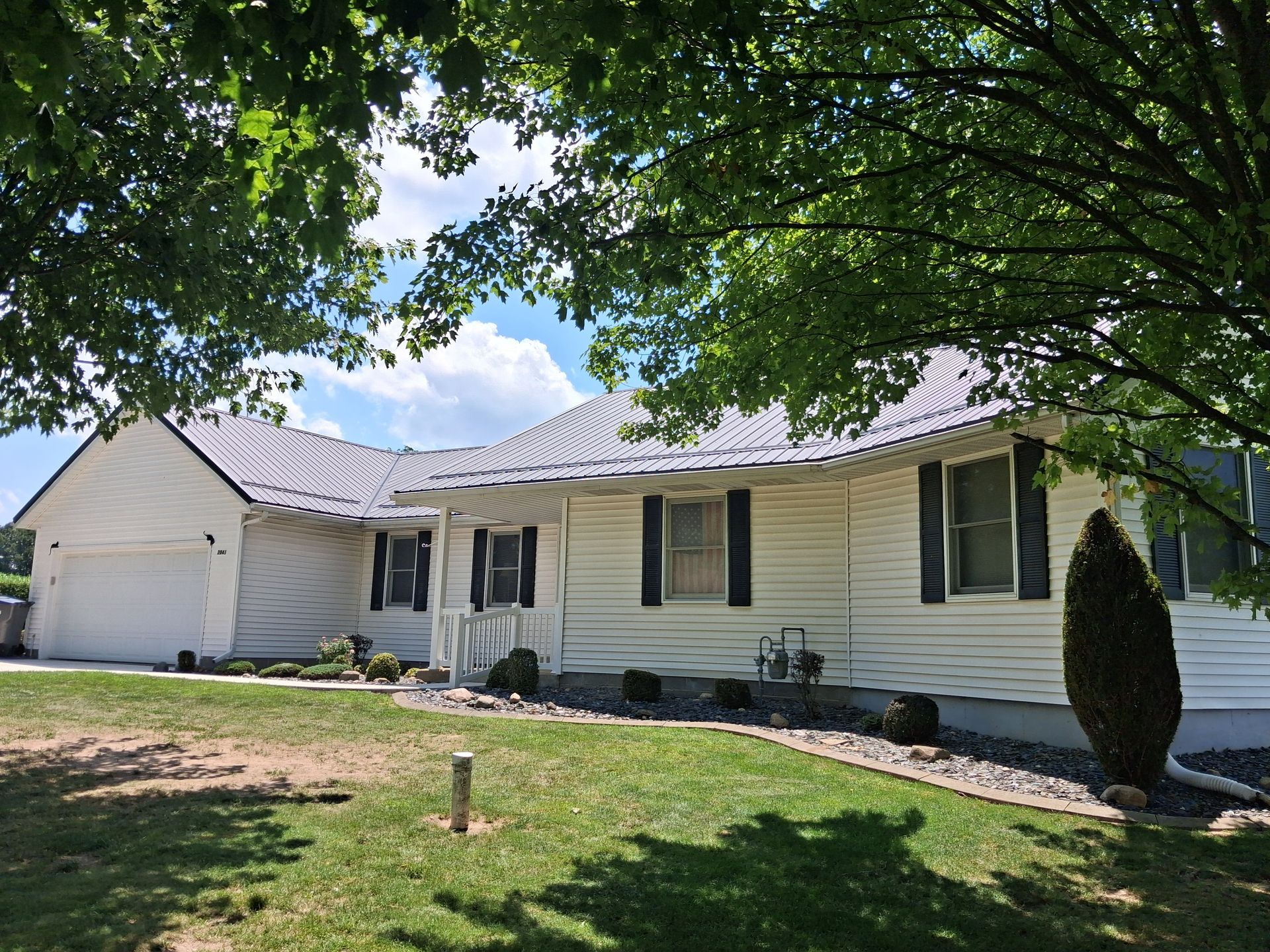 White single-story house with garage, black shutters, and a grassy yard under leafy trees