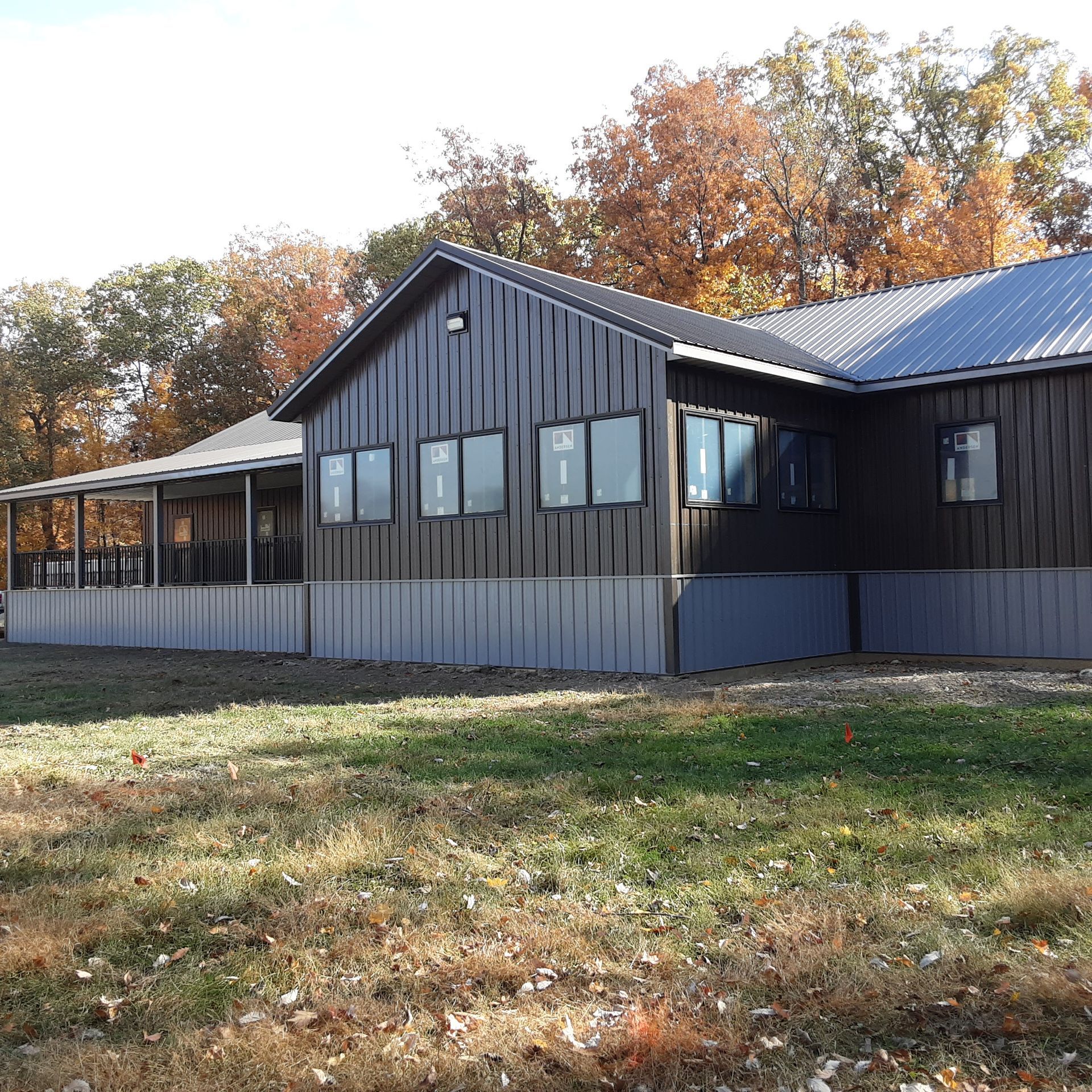 Dark gray building with metal roof and covered porch, surrounded by autumn trees and grassy yard.