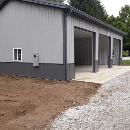 Gray metal garage building with open bays, concrete apron, and gravel driveway