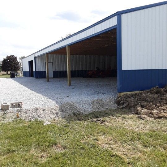 Blue-and-white metal barn with open bays and gravel lot beside a grassy field