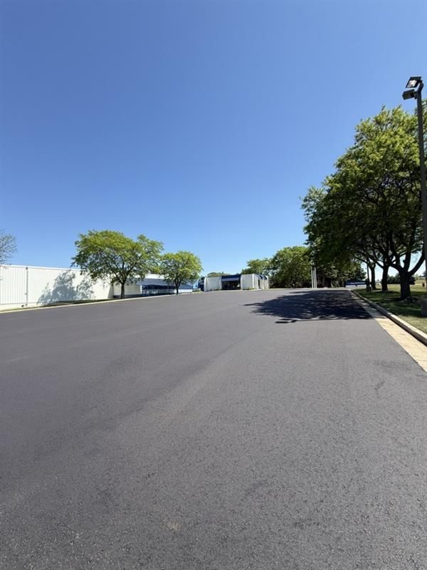 Wide paved road beside trees and a white wall under a clear blue sky.