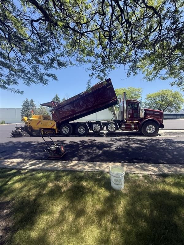 Red dump truck tipping a maroon trailer into a yellow loader on a sunny roadside