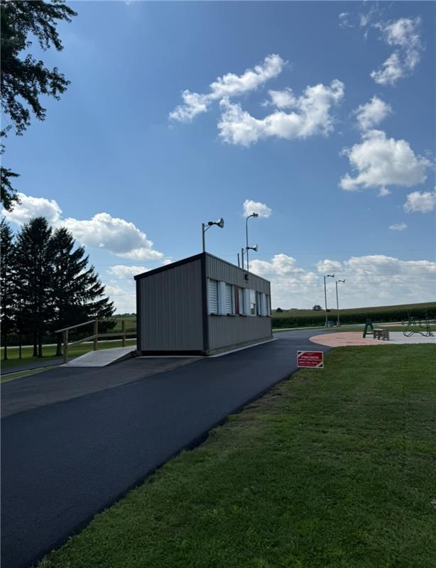 A small gray building with a ramp sits on a paved path, on a grassy area, under a blue sky.