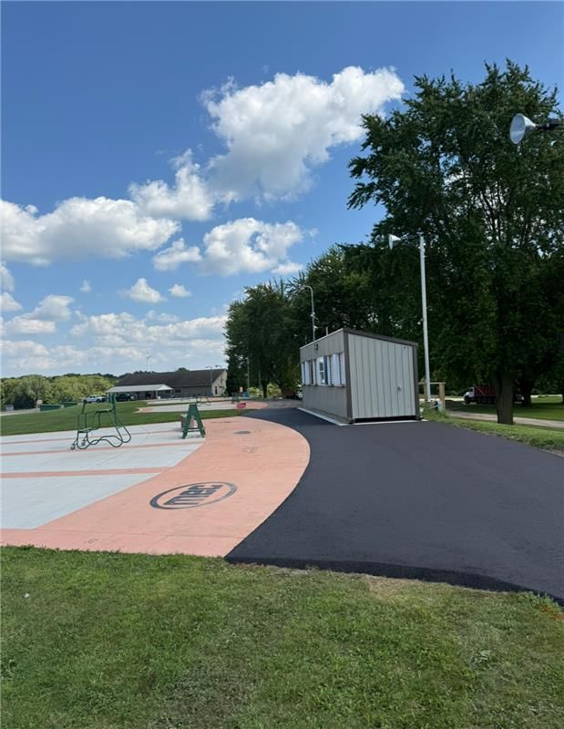 Asphalt path curves past a small building with light-colored siding in a park on a sunny day.