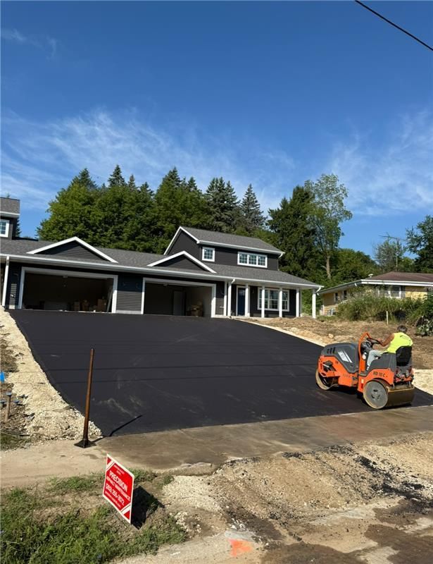 Asphalt paving in progress on a driveway leading to a gray house; roller compacting surface; blue sky.