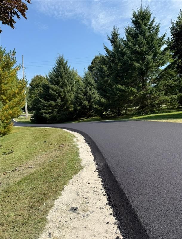 Newly paved asphalt road curves through a grassy area, with trees in the background under a clear blue sky.