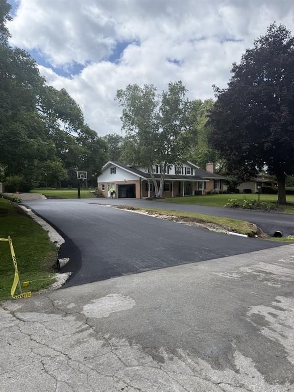 Newly paved driveway leading to a house with a garage and porch, trees on either side, cloudy sky.