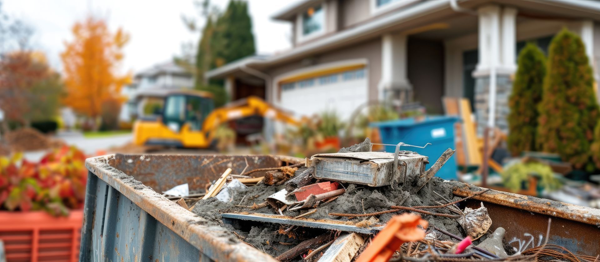 A close-up of a full dumpster with debris, a house, and an excavator in the background.