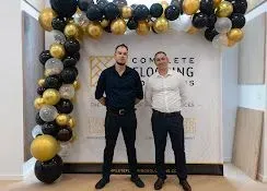 Two men standing under a black, gold, and silver balloon arch at a clothing event backdrop