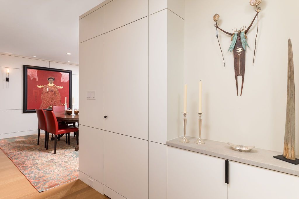 Modern hallway with white cabinets, minimalist decor, and a dining room with red chairs and a large red artwork.