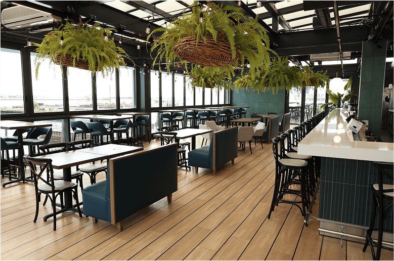 Modern restaurant interior with hanging plants, wooden floors, and rows of black tables and chairs
