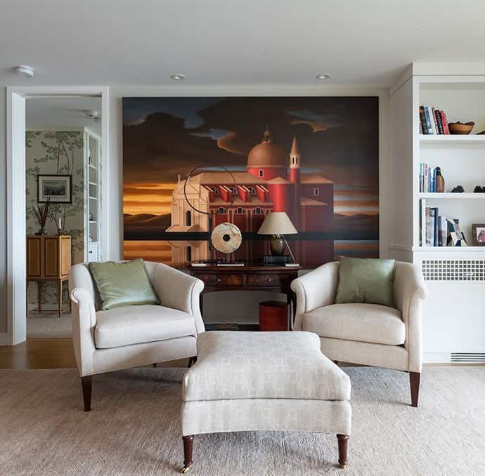 Modern living room with two beige armchairs, an ottoman, and a large wall mural behind a side table and lamp.