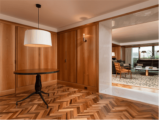 Modern wood-paneled room with herringbone floor, round table, pendant lamp, and view into a bright living area.