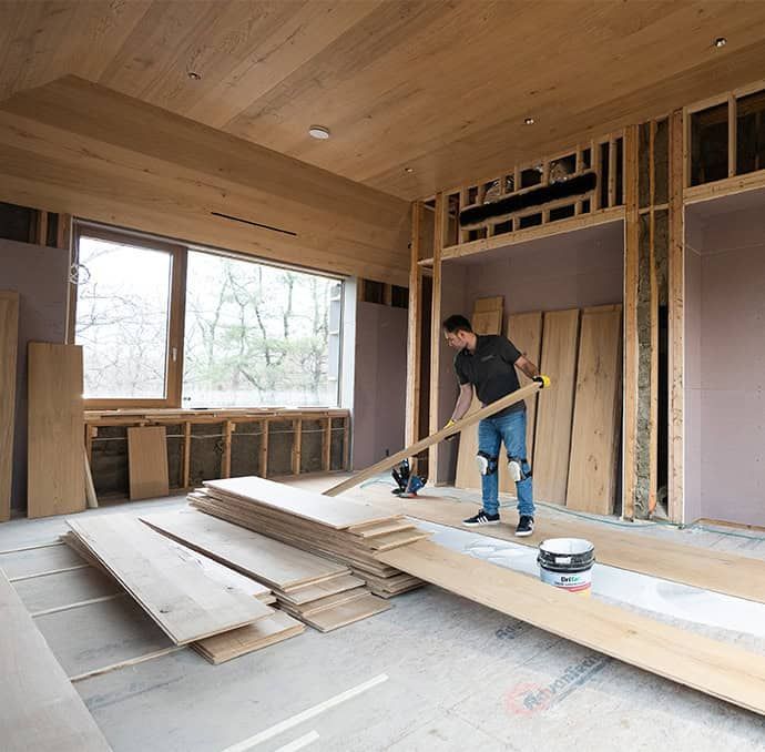 Worker sweeping wood planks in an unfinished room with exposed framing and windows.