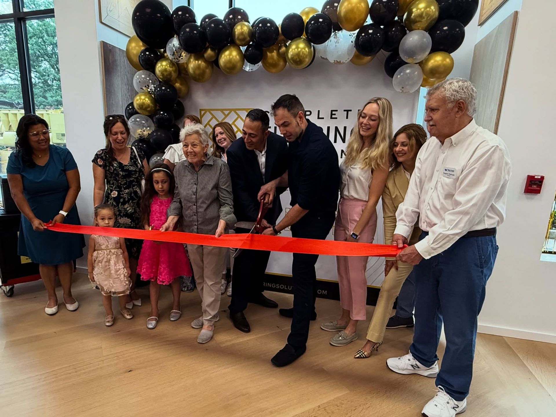 Ribbon-cutting ceremony with people holding a red ribbon under black, gold, and white balloons in a bright room