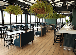 Modern restaurant dining area with wood floor, black chairs, hanging plants, and large windows
