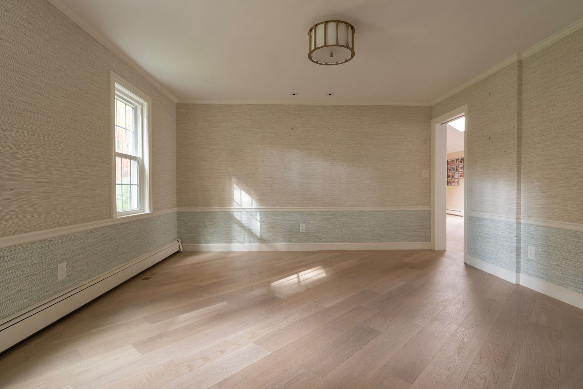Empty sunlit room with light wood floors, pale walls, and a window on the left.