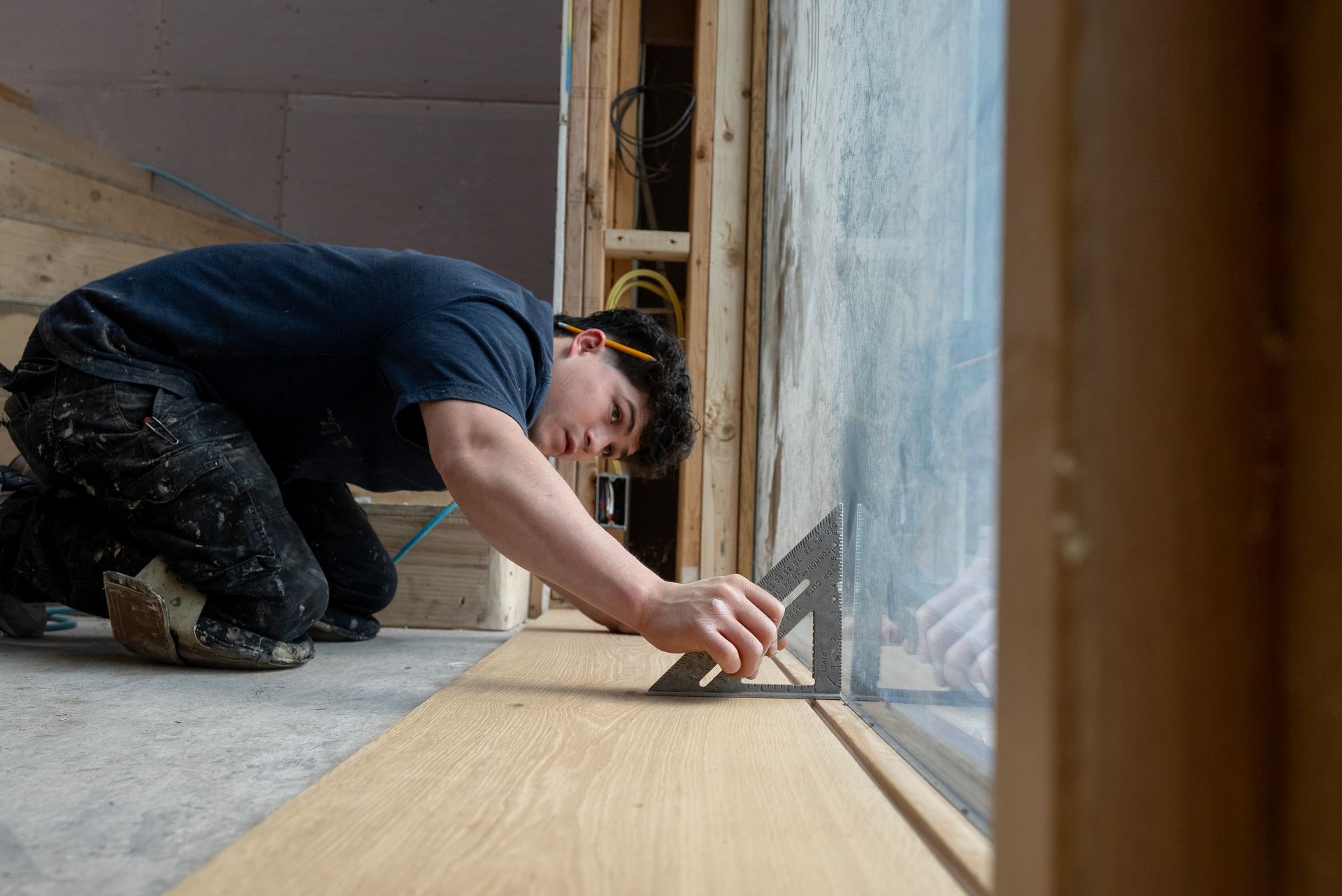 Worker kneeling on a floor, carefully installing wood flooring beside a wall with a tool.