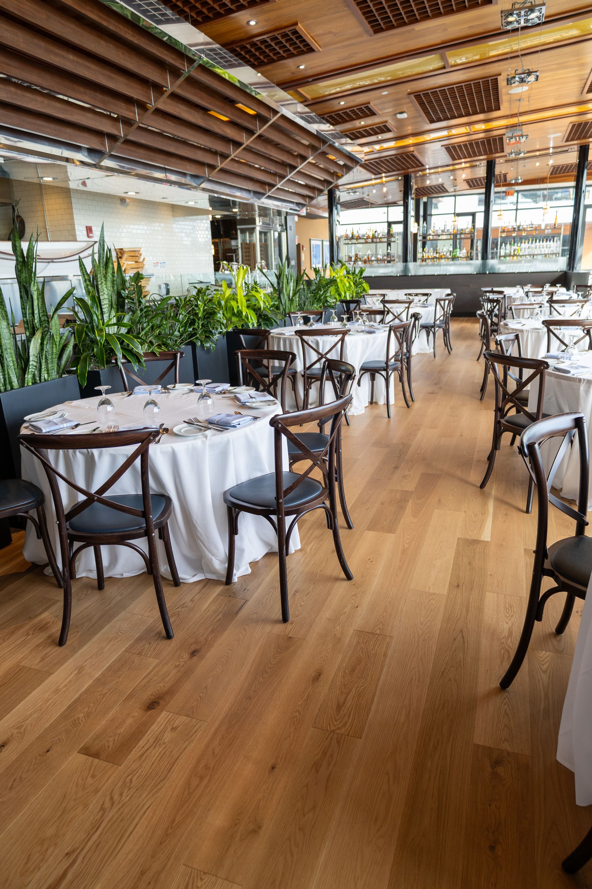 Empty restaurant dining room with wooden floors, white tablecloths, and plants by large windows