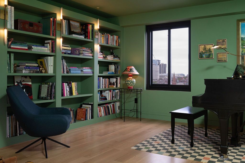 Green room with bookshelves, blue chair, piano, and a window overlooking buildings