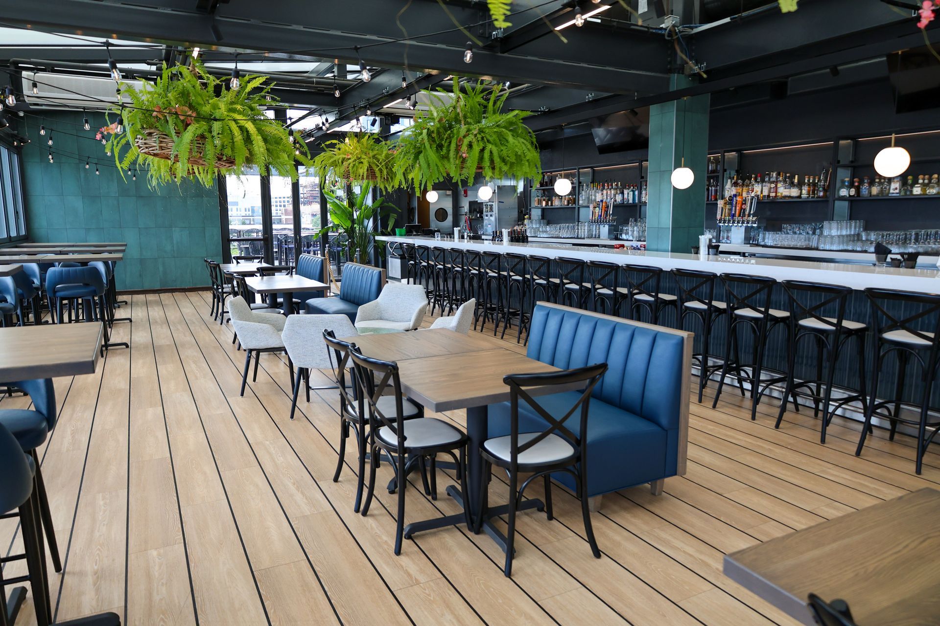 Modern restaurant patio with wooden deck, blue banquette seating, hanging plants, and bar stools