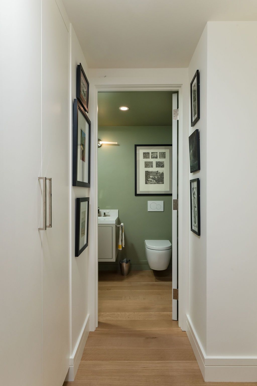 Narrow hallway with white cabinets opening to a green bathroom with a toilet visible.