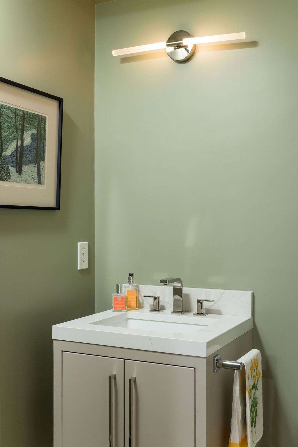 Small bathroom vanity with white sink, beige cabinet, wall light, and framed art on green wall.