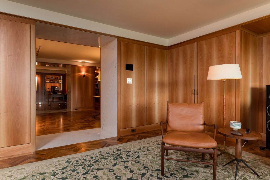 Wood-paneled lounge with two leather chairs, lamp, patterned rug, and a doorway to a warmly lit hall.