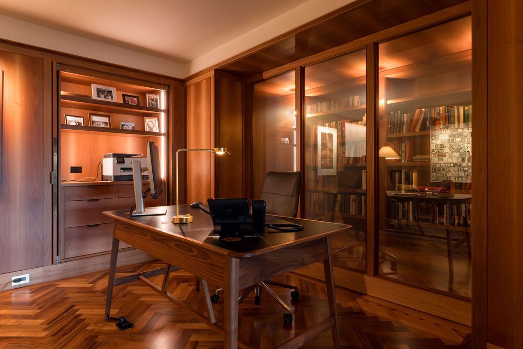 Wood-paneled office with a desk, chairs, and large glass cabinets reflecting the warmly lit room