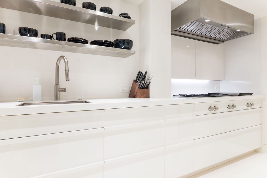 Bright white modern kitchen with long counters, open shelves, sink, and stainless steel range hood