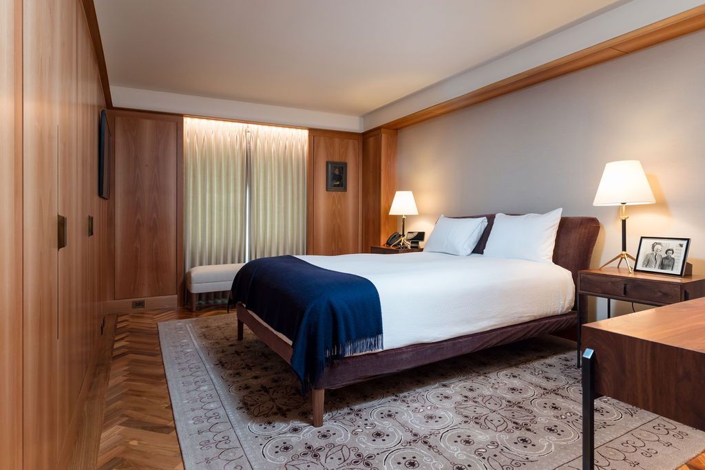 Hotel bedroom with a neatly made bed, blue throw blanket, bedside lamps, and wood-paneled walls.