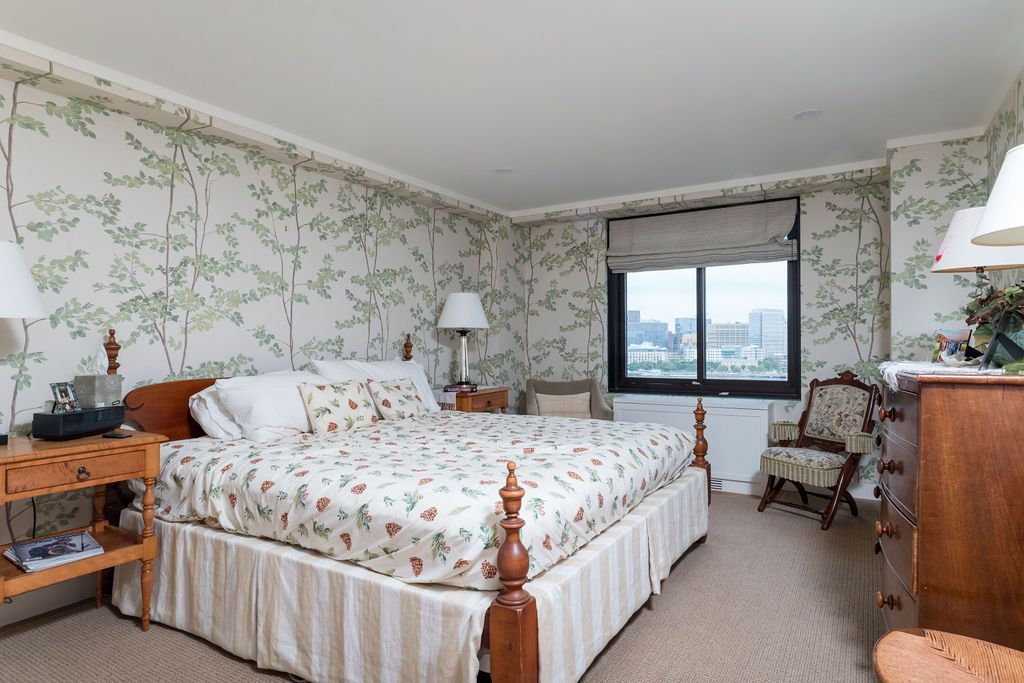 Bedroom with floral wallpaper, wooden bed, armchair, and large window overlooking a city view