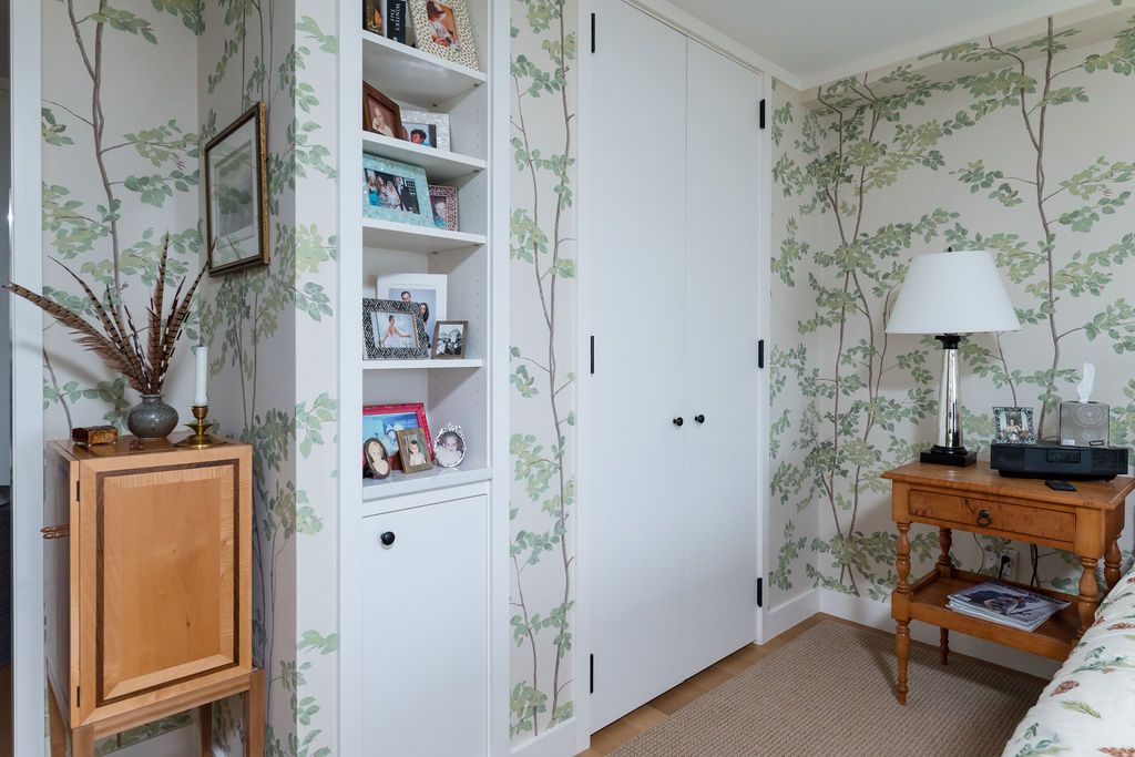 Bright bedroom corner with floral wallpaper, white closet doors, shelves, and a wood side table with lamp.