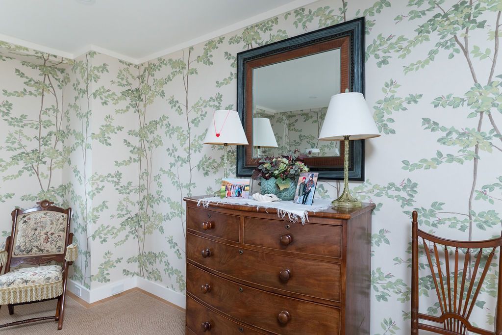 Floral wallpaper bedroom corner with a wooden dresser, mirror, lamps, and a chair