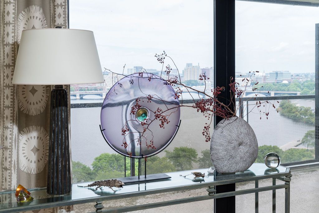 Decorative floral arrangement with a circular sculpture on a glass table by a large window overlooking water