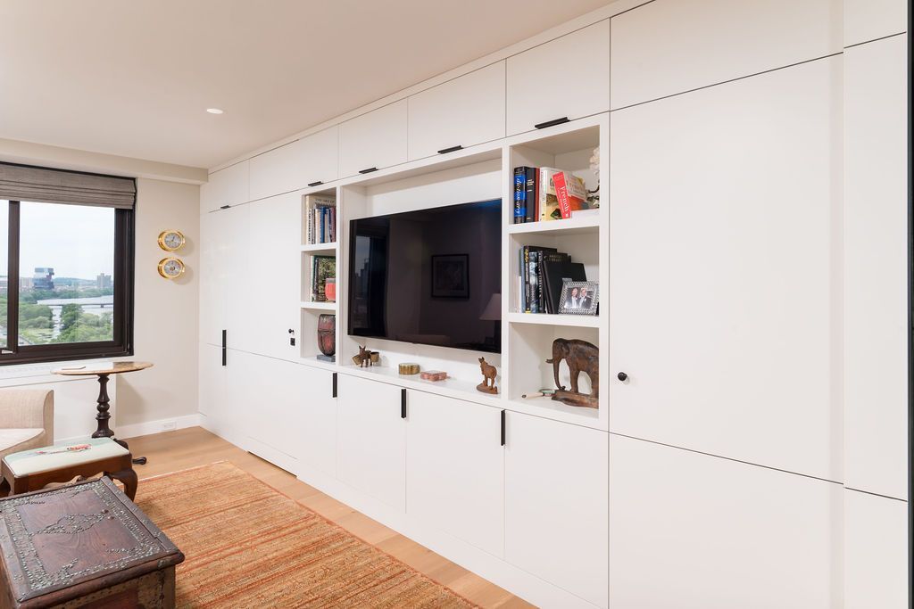 Bright living room wall with built-in white cabinets, a TV, shelves, a window, and a woven bench near a rug.