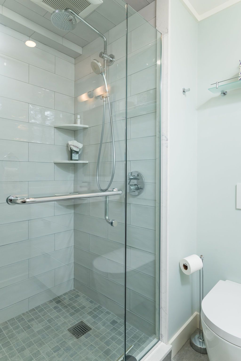 Modern bathroom with glass walk-in shower, tiled walls, chrome grab bars, and toilet edge visible