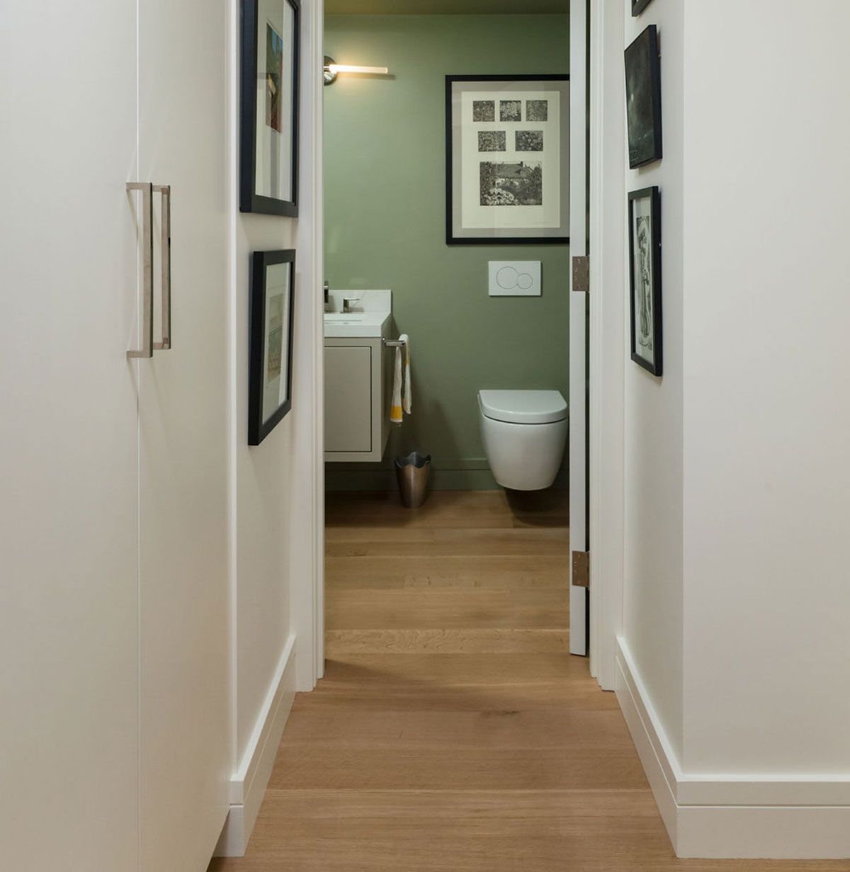Hallway view into a small bathroom with green walls, wood floor, and white toilet under framed pictures.