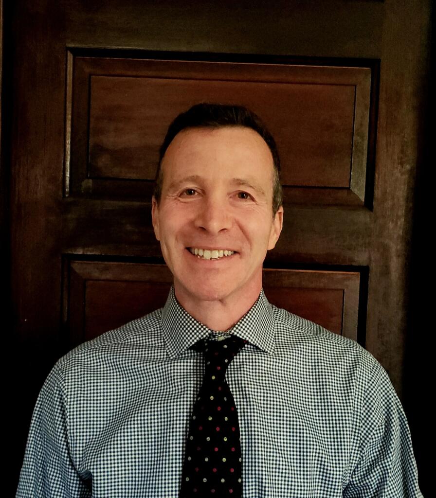 Brian Richards stands in front of a wooden door and smiles at the camera.