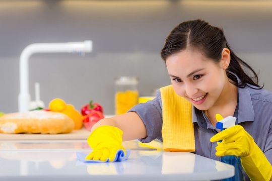 Woman cleaning kitchen countertop with yellow gloves and spray bottle.