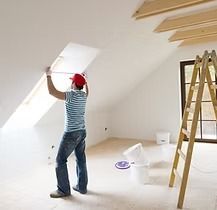 Man in a red hat measures a white angled ceiling in a bright room. A ladder and buckets are nearby.